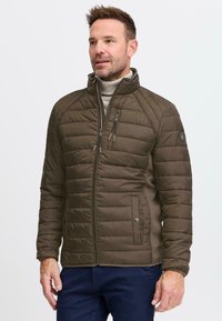 Mann iført olivengrønn quiltet jakke og lys grå turtleneck, står mot en ensfarget hvit bakgrunn.