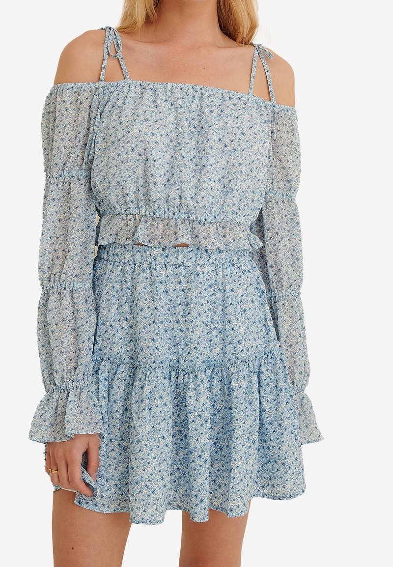 Donna che indossa un vestito off-shoulder floreale azzurro chiaro con maniche lunghe a sbuffo e orlo arricciato, mentre tiene delicatamente la gonna con una mano.