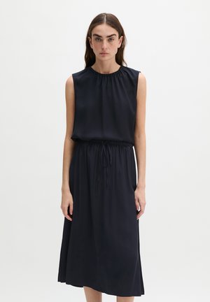 Femme aux longs cheveux bruns portant une robe midi noire sans manches avec un col froncé et une taille à cordon, debout sur un fond uni.