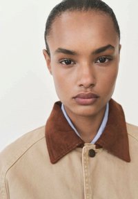 Beige jacket with a brown corduroy collar, featuring a button closure. The fabric is soft, with a structured design and contrasting collar texture.