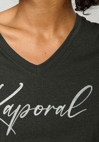 T-shirt vert foncé en V en coton doux, avec un texte script argenté brillant au design fluide. Le texte indique "Kaporal."