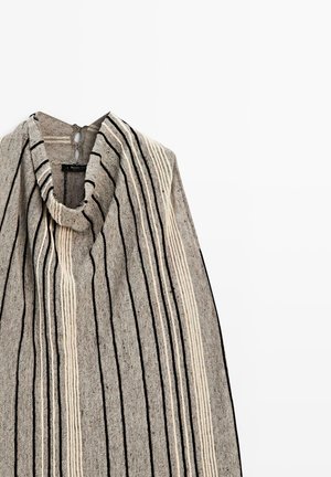 Poncho gris tricoté sans manches avec des rayures verticales noires et blanches et un col bénitier drapé suspendu sur un fond blanc.