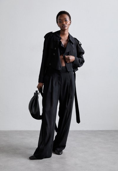 Črn skrajšan plašč s širokim ovratnikom in gumbi, v kombinaciji z ohlapnimi črnimi hlačami. Dodatki vključujejo črno torbico in ravne čevlje.