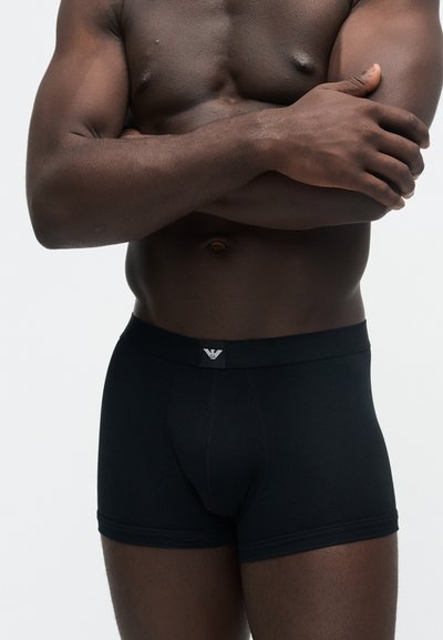 Homme musclé à la peau foncée portant un boxer noir avec un petit logo sur la ceinture, les bras croisés sur sa poitrine nue.
