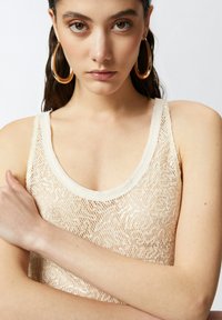 Bež majica bez rukava ukrašena šljokicama s izrezom u obliku u obliku slova U i širokim trakama. Tkanina ima mrežastu teksturu, a boja je meka i neutralna.