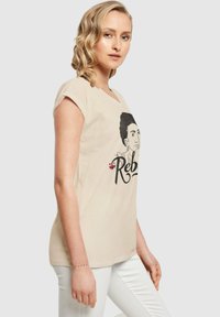 Beige Baumwoll-T-Shirt mit kurzen Ärmeln, das eine grafische Illustration und das Wort "Rebel" in schwarzem Text zeigt. Leicht und locker geschnitten.