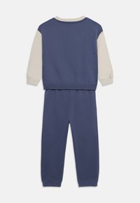 Zweiteiliges Trainingsanzug in Marineblau und Creme. Langärmliges Oberteil mit Rundhalsausschnitt und elastischen Bündchen, passende Hose mit elastischem Bund und Bündchen.