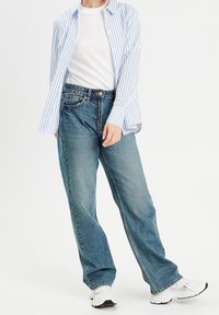 Lichtblauw en wit gestreepte button-up shirt over een wit T-shirt, gecombineerd met losse blauwe denim jeans en witte sportschoenen.