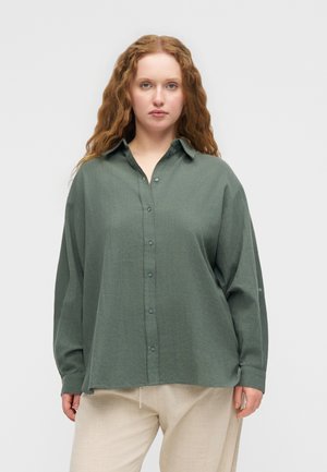 Kvinde med langt krøllet rødt hår iført en løs grøn skjorte med knapper og lyse beige bukser, stående foran en ensfarvet lys baggrund.