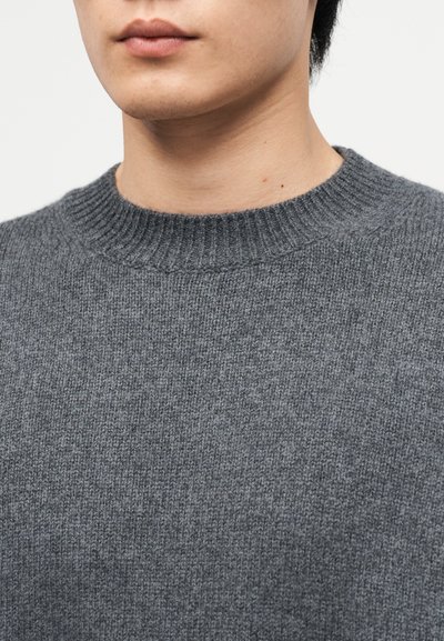 Gros plan sur la partie inférieure du visage et le cou portant un pull à col rond en maille grise texturée, sur fond uni.