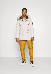 Parka chiaro con dettagli in pelliccia sintetica sul cappuccio, accenti neri, dettagli con bottoni, abbinata a pantaloni cargo gialli e calzature sportive.
