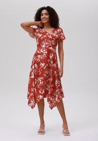 Robe rouge avec un motif floral blanc, manches courtes, encolure en V et ourlet asymétrique. Tissu lisse, design léger et longueur jusqu'aux chevilles.