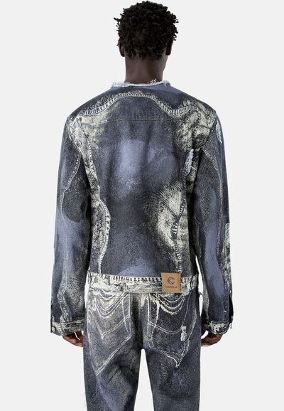 Personne portant une veste en denim noir et blanc délavée à l'acide et un jean assorti, debout dos à la caméra sur un fond uni.