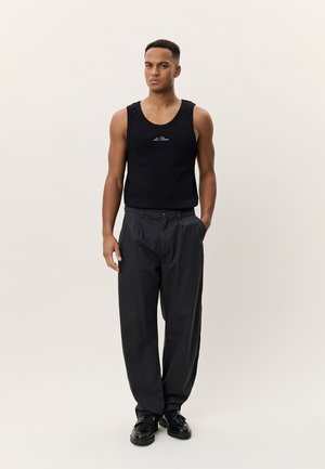 Νεαρός άνδρας όρθιος, φοράει μαύρο αμάνικο μπλουζάκι με λογότυπο, φαρδιά σκουρόχρωμη παντελόνα και μαύρα παπούτσια με κορδόνια, ουδέτερο φόντο.