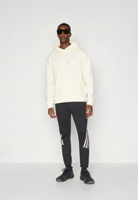 Λευκό φούτερ με κουκούλα και ριμπ μανσέτες, συνδυασμένο με μαύρο στενό παντελόνι φόρμας με λευκές ρίγες στα πλαϊνά. Το μοντέλο φοράει μαύρα αθλητικά παπούτσια.