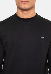 Sweat-shirt noir en coton, avec un col ras du cou, des poignets côtelés et un petit écusson logo en forme de diamant sur le côté gauche de la poitrine. Texture lisse.