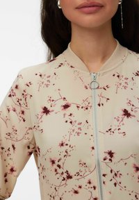 Femme portant une veste beige à fleurs roses et rouges foncées, dotée d'une fermeture éclair argentée avec une languette ronde.