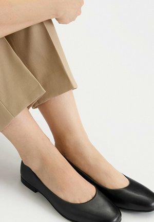 Pies con bailarinas de cuero negro combinadas con pantalones beige recortados sobre un fondo liso y claro.