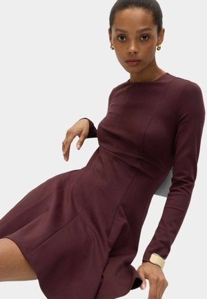 Kvinde med guldbøjler og armbånd iført en figursyet bordeauxrød langærmet kjole, siddende mod en lys baggrund.