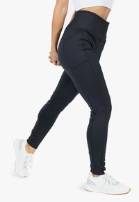 Leggings sportivi neri con vita alta, dotati di tasche laterali, tessuto morbido e dettagli di cucitura a contrasto. Indossati con sneakers bianche.
