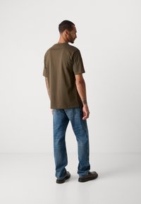 Bărbat cu tatuaje îmbrăcat într-un tricou verde măsliniu, blugi albaștri și pantofi maronii, stând cu spatele într-un fundal alb simplu.