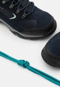Gros plan sur des chaussures de randonnée bleu marine avec des semelles noires et des œillets métalliques, à côté d'un lacet vert sarcelle noué solidement.