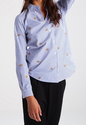 Femme portant une chemise à rayures bleues et blanches avec de petits motifs brodés en forme d'yeux, associée à un pantalon noir, posant avec un bras levé.
