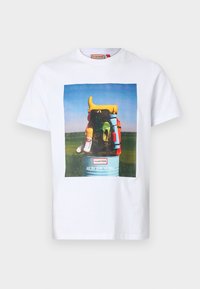White cotton t-shirt featuring a colorful graphic of boots and bags against a blue sky, with text reading "All the gear, no idea."