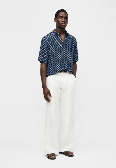 Moški stoji oblečen v temno modro srajco s kratkimi rokavi z vzorcem, bele široke hlače, črni pas in črne sandale na enobarvnem ozadju.