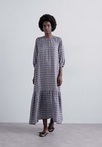 Rochie midi cu model mov, cu mâneci bufante, hem strâns și material ușor. Purtați-o cu sandale plate negre.