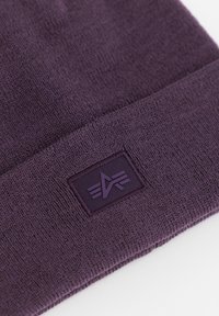 Ljubičasti pleteni šešir s preklopom koji ima strukturiranu, kvadratnu naljepnicu. Naljepnica prikazuje logotip u malo tamnijoj nijansi ljubičaste.