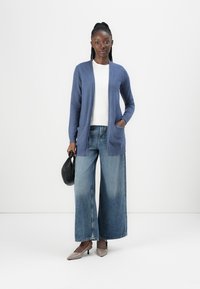 Blå cardigan med långa ärmar, två framfickor, bärs över en vit topp, i kombination med vida denimjeans och grå klackar. Svart handväska ingår.