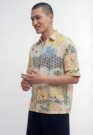 Młody mężczyzna z krótkimi włosami, ubraną w kolorową wzorzystą koszulę z krótkim rękawem i ciemne spodnie, uśmiechający się i gestykulujący rękami.