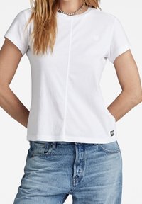 T-shirt en coton blanc à manches courtes, présentant un design minimaliste et un logo discret sur le devant. Assorti à un jean en denim bleu clair.