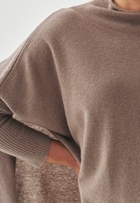 Długi rękaw, jasnobrązowy sweter dzianinowy o luźnym kroju, z szerokim dekoltem i prążkowanymi mankietami, które dodają tekstury. Miękki, gładki materiał.