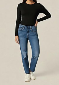 Femme portant un haut noir à manches longues, un jean bleu droit et des bottines blanches, debout avec une main sur la hanche.