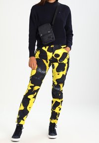Svarta och gula mönstrade byxor med en djärv splatterdesign. I sällskap med en mörk stickad tröja och en liten svart axelväska.