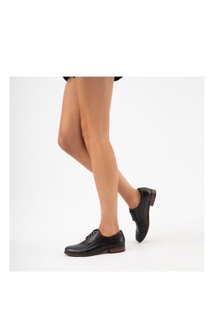 Personne portant des chaussures Oxford en cuir noir à lacets avec semelles marron, montrant les jambes de la mi-cuisse aux pieds sur un fond blanc.