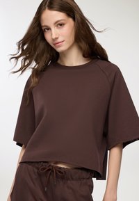 Top brown over-size a maniche corte realizzato in un tessuto testurizzato con scollatura rotonda. Orlo cropped notevole e vestibilità rilassata. Visibili pantaloni abbinati con Coulisse.