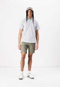 Chemise à manches courtes à rayures blanche et bleue, associée à un short olive. Le mannequin porte une casquette gris clair et des baskets blanches avec des accents noirs.