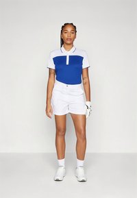 Polo azul e branco com mangas curtas e uma pala frontal, combinado com calções brancos e uma luva de golfe. Ténis brancos completam o conjunto.