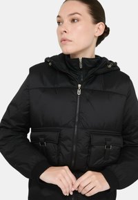 Giacca a vento nera con cappuccio aderente, zip frontale e due tasche con pattina. Presenta polsini a coste e una texture morbida e trapuntata.