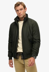 Dunkelgrüne, mit Reißverschluss versehenen Jacke mit hohem Kragen; verfügt über Seitentaschen, glatte Textur und subtile Streifen. Wird über einem cremfarbenen Pullover getragen.