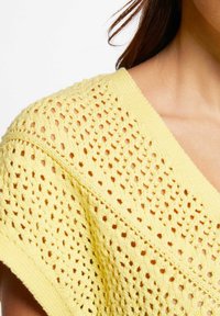 Maglione giallo lavorato a maglia con un design a traforo, dotato di scollatura a V e maniche corte. La texture è traspirante con una vestibilità ampia.