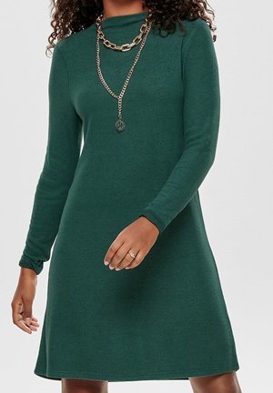 Woman wearing a long-sleeve green dress with two layered gold chain necklaces and a ring on her finger.