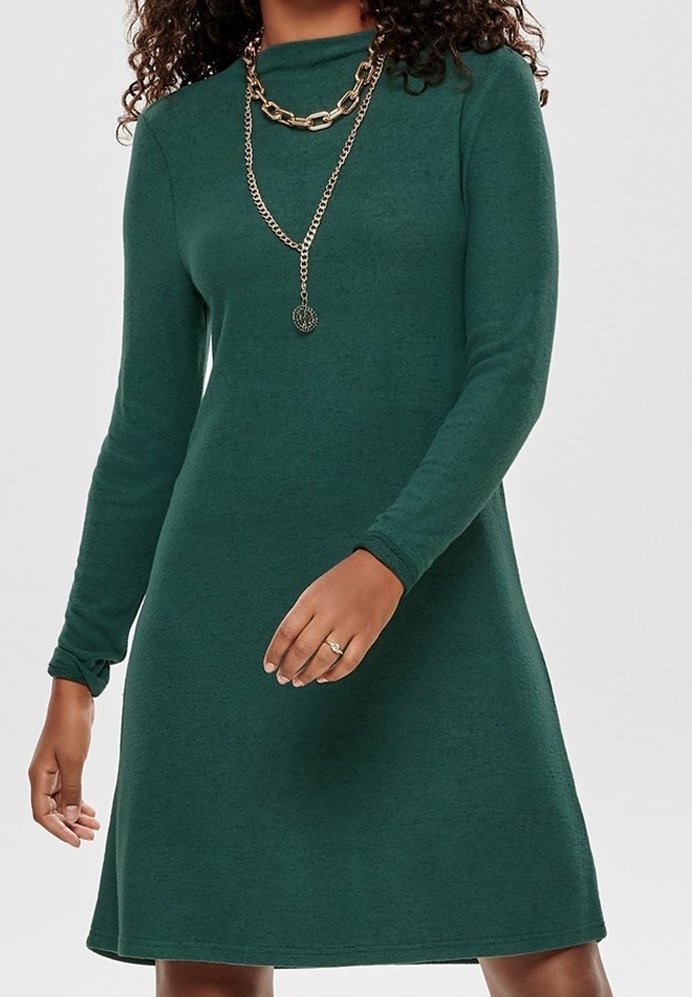 Femme portant une robe verte à manches longues avec deux colliers en chaîne dorée superposés et une bague à son doigt.