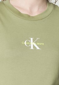 T-shirt en coton vert olive avec un col rond, arborant le logo "Calvin Klein Jeans" en blanc et jaune imprimé au centre de la poitrine.