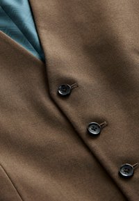 Vesta din material maro cu un finisaj texturat, prezentând trei nasturi negri și o căptușeală teal cu un model geometric.