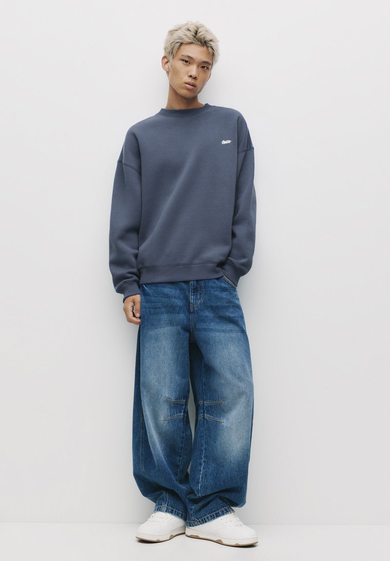 Tengerészkék, oversized pulóver bordás mandzsettával, világoskék, bő szabású farmernadrággal, látható varrásokkal és laza sziluettel, fehér tornacipőkkel párosítva.