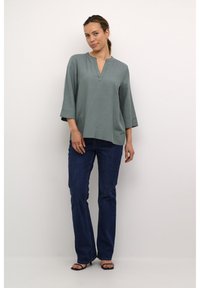 Top verde chiaro a tre quarti con scollatura a V, abbinato a jeans fitted blu scuro. Tocco liscio, vestibilità casual.